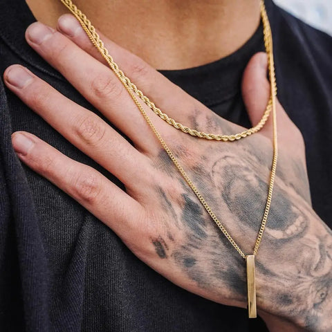Man layering two gold chains and a bar pendant on a crew-neck tee — how to layer necklaces men